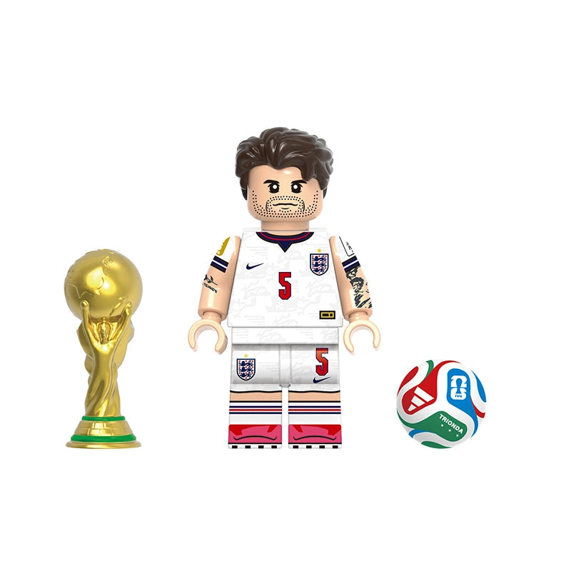 A toy soccer minifigure with a white England jersey, number 5, standing next to a gold FIFA trophy and a multicolor soccer ball on a white background. A gray label at the top reads 'GH0821' and 'Stones' is printed below.